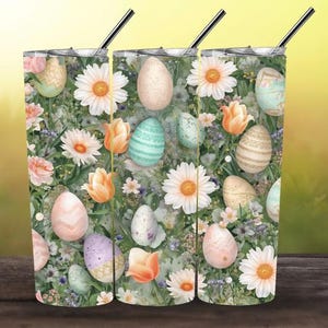 May include: Three stainless steel tumblers with black straws. The tumblers feature a floral design with Easter eggs, daisies, and tulips. The eggs are pastel-colored and decorated with patterns. The background is a mix of green and white.