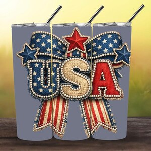 May include: A gray cylindrical tumbler with a straw featuring a graphic of the letters USA in a burlap texture, surrounded by a blue denim bow with white stars, a red star, and red and white striped ribbon accents with a pearl-like border.