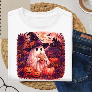 May include: A white t-shirt featuring a graphic print of a ghost wearing a witch hat and holding a pumpkin-themed beverage. The scene includes a haunted house, bats, a crescent moon, orange pumpkins, and purple roses in a watercolor style.