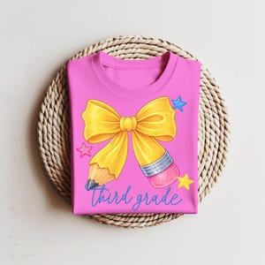 Puede incluir: Una camiseta rosa brillante con un gráfico de un lápiz amarillo con forma de lazo. El lápiz tiene una virola plateada y una goma de borrar rosa. El texto "third grade" está impreso en cursiva azul debajo del lazo, junto a pequeñas estrellas azules y amarillas.