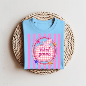 Könnte beinhalten: Ein hellblaues T-Shirt mit vertikalen rosa Streifen und einer zentralen Grafik eines rosa-weiß karierten Apfels. Der Apfel ist von einem gewellten kreisförmigen Rahmen mit kleinen bunten Sternen und dem Schriftzug third grade umgeben.