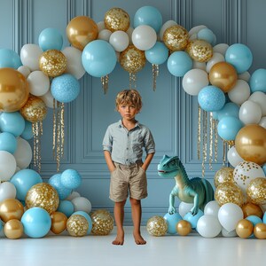May include: A blue and gold balloon arch with a young boy standing in front of it. The boy is wearing a light blue shirt and khaki shorts. A green dinosaur toy is in front of the boy.