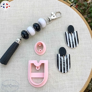 Puede incluir: Un llavero hecho a mano con cuentas blancas y negras, una borla negra y un cierre plateado. También se muestran dos pendientes a rayas blancas y negras y dos cortadores de arcilla polimérica rosas.