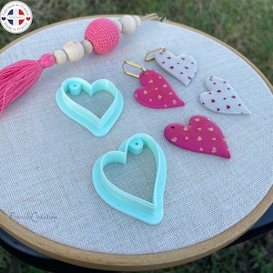May include: An assortment of heart-shaped craft supplies. Two light blue heart-shaped cookie cutters and three pink and white heart-shaped charms with gold earring hooks are arranged on a fabric surface. A pink tassel and wooden beads are also visible.