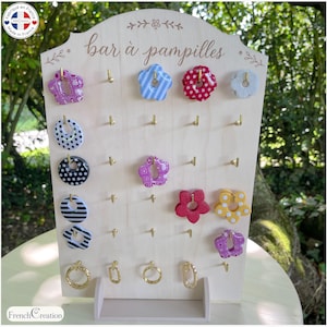Peut inclure: Un présentoir à bijoux en bois clair avec le texte bar à pampilles gravé en haut. Le support comporte plusieurs crochets en métal doré retenant divers charms colorés, à motifs et de formes variées, notamment des fleurs, des cercles et des anneaux.