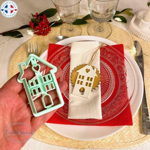 May include: A light green house-shaped cookie cutter with a heart detail is held near a place setting. A wooden house-shaped tag with a heart cutout rests on a white napkin. The place setting includes a red plate and a clear glass plate.