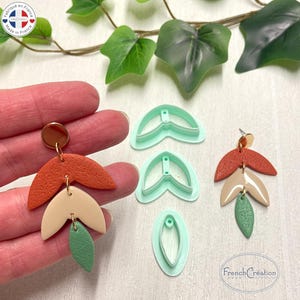 May include: Handmade orange, beige, and green leaf-shaped earrings with gold-coloured hardware. The earrings are displayed with matching earring cutters. A unique accessory.