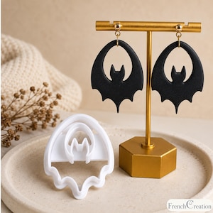 May include: A pair of matte black bat-shaped dangle earrings hanging from a gold-toned T-bar display stand, accompanied by a white plastic bat-shaped cookie cutter. The earrings feature a cutout design with a central bat silhouette.