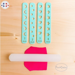 Peut inclure: Quatre outils de modelage turquoise avec des découpes en forme de croissant de lune, de losange, de cœur et d'étoile, chacun mesurant en mm. Un rouleau à pâtisserie blanc repose sur de l'argile rose. Sur une surface en bois.