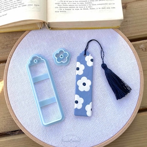 Puede incluir: Un marcapáginas azul claro con diseños de flores blancas y una borla azul marino, junto con un cortador de manualidades en forma de flor y un cortador en forma de marcapáginas. Los artículos están sobre tela blanca.
