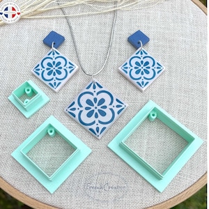 May include: A set of four blue and white ceramic tile-shaped cookie cutters. The cutters are in different sizes and feature a floral design. The cutters are on a light brown surface.