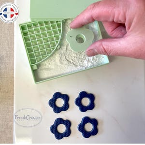Puede incluir: Un molde de plástico verde con un patrón de rejilla y un recorte en forma de flor. Una mano sostiene un molde de plástico verde en forma de flor. Cuatro moldes en forma de flor azules están sobre una superficie blanca. El texto "French Création" es visible en la superficie.