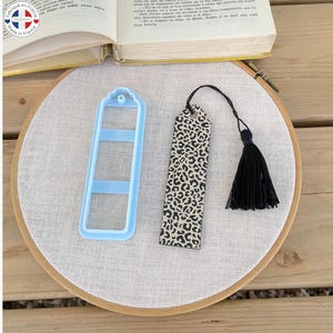 Puede incluir: Una herramienta de manualidades azul claro con forma de marcapáginas y un marcapáginas con estampado de leopardo con una borla negra descansan sobre un bastidor de bordado cubierto de tela blanca. Un libro abierto está en el fondo.