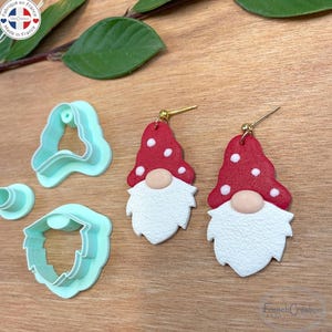 Peut inclure: Boucles d'oreilles gnome faites à la main avec des chapeaux rouges et des barbes blanches, ainsi que des emporte-pièces en pâte polymère bleu clair. Les boucles d'oreilles ont des crochets dorés. L'image est sur une surface en bois.