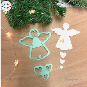 Könnte beinhalten: Auf einer Holzoberfläche werden Ausstecher und Ausschnitte in Engelsform präsentiert, darunter ein hellgrüner Engels-Ausstecher, ein weißer Engels-Ausschnitt mit einem gelben Herzen und weiße Herz-Ausschnitte. Eine Lichterkette in Sternform und Tannenzweige sind ebenfalls vorhanden.