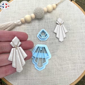 Könnte beinhalten: Handgefertigte, hellfarbene Ohrringe mit geometrischem Art-Deco-Design, gehalten in einer Hand, neben zwei weiteren Ohrringen und zwei blauen Ohrringausstechern. Ein Perlenquasten und ein Aufkleber Made in France sind im Hintergrund.