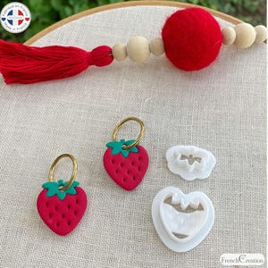 Peut inclure: Deux breloques en forme de fraise avec des sommets verts et des corps rouges, chacune attachée à un anneau doré. Sont inclus deux emporte-pièces en plastique blanc, un en forme de fraise et un en forme de fleur, ainsi qu'un pompon rouge avec des perles en bois.