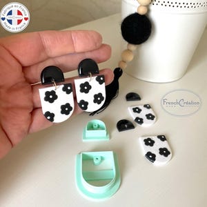 May include: Handmade earrings featuring a black and white floral pattern. The earrings have a black upper section and a white base adorned with black flower motifs. The image also shows earring components and a cutting tool.
