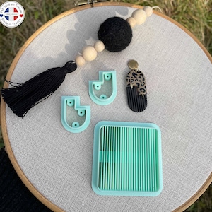 Könnte beinhalten: Eine Auswahl an Bastelwerkzeugen und Schmuckkomponenten ist auf einer neutralen Oberfläche angeordnet. Die Artikel umfassen hellblaue Polymer Clay Cutter, einen Ohrring mit Leopardenmuster und Schwarz sowie ein quadratisches Werkzeug. Ein schwarzer Quasten- und Holzperlen-Girlande sind ebenfalls vorhanden.
