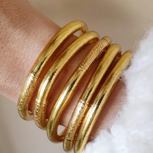 Op de afbeelding: Vier gouden armbanden gestapeld op een pols. De armbanden zijn gemaakt van een glanzend, metalen materiaal en hebben een licht getextureerd oppervlak.