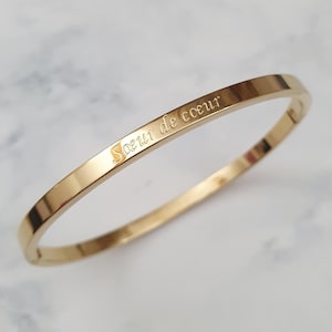 Puede incluir: Una pulsera dorada con la inscripción "Soeur de coeur" grabada en la parte superior. La pulsera tiene un diseño sencillo y elegante y un acabado pulido. Es una pieza de joyería.