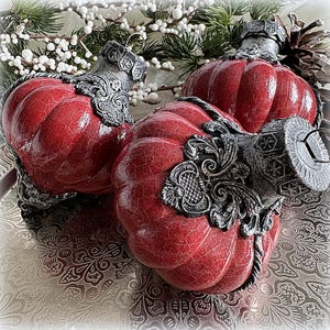 Könnte beinhalten: Drei rote, kürbisförmige Ornamente mit glänzender Rissoptik. Jedes Ornament hat eine dunkle, verzierte Kappe mit metallischer Textur, komplizierten Schnörkeln und einem Sechseckmuster. Die Ornamente sind abgerundet und weisen vertikale Rippen auf.