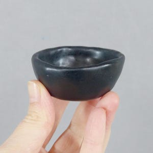 Puede incluir: Un pequeño cuenco de cerámica negra con un borde ligeramente irregular. El cuenco se sostiene en una mano sobre un fondo gris neutro. El color oscuro y el diseño simple sugieren una estética minimalista, adecuada para servir pequeñas porciones.