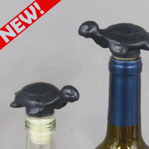 Può includere: Due tappi per bottiglia neri a forma di tartaruga. Uno è su una bottiglia trasparente, l'altro su una bottiglia di vino con etichetta blu. La parola "NEW!" è stampata in bianco su uno striscione diagonale rosso in alto a sinistra.