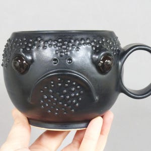 May include: Black ceramic mug with a textured surface and a frog face design. The mug has a large handle and two black eyes.