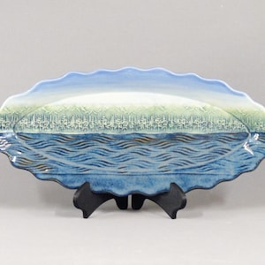 May include: An oval ceramic serving platter with a scalloped edge featuring a landscape illustration. The design depicts a blue sky, a pale green mountain range, a forest of white pine trees, and a textured dark blue body of water with waves.