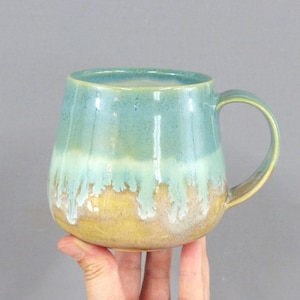 May include: A ceramic mug featuring a rounded body and a curved handle. The upper portion is a deep teal color, transitioning into a lighter mint green drip pattern that overlays a warm, earthy tan base at the bottom of the vessel.