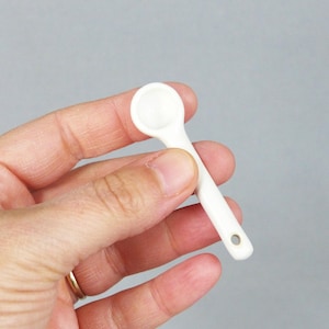 May include: A small, white ceramic spoon with a rounded bowl and a long handle. The handle has a small hole at the end. The spoon is held in a hand against a gray background. This spoon is likely for measuring small quantities of ingredients.
