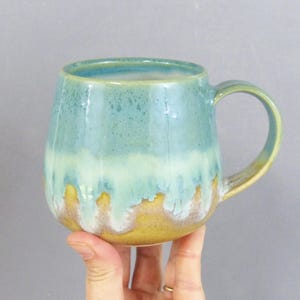 May include: A ceramic mug featuring a rounded body and a curved handle. The exterior displays a gradient glaze, transitioning from a deep teal at the rim to a lighter mint green in the center, and finishing with a warm mustard yellow at the base.