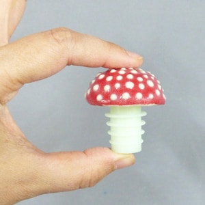 Può includere: Un tappo per bottiglia a forma di fungo rosso e bianco. Il cappello del fungo è rosso con pois bianchi. Lo stelo è di colore verde chiaro. Il tappo è tenuto in mano su sfondo grigio.