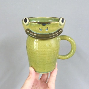 May include: A handmade ceramic mug shaped like a frog. The mug is predominantly light green with darker green and brown details. The frog's eyes and mouth are detailed, and the handle is a simple loop.