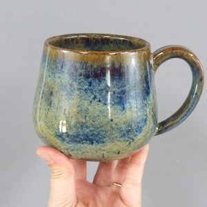May include: A ceramic mug with a rounded body and a curved handle, featuring a reactive glaze finish in mottled shades of deep blue, earthy green, and warm brown tones that blend together across the textured surface of the vessel.
