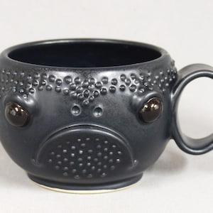 May include: Black ceramic mug with a textured surface and a cartoon face with large eyes. The mug has a handle.