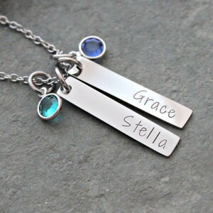 May include: A silver necklace with two silver bars engraved with the names "Stella" and "Grace". The necklace has two small blue and green gemstones.