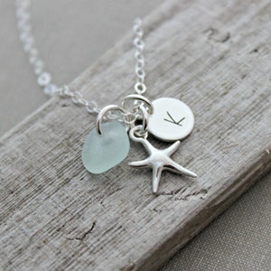 May include: A silver necklace with a light blue sea glass charm, a silver starfish charm, and a silver disc charm with the letter "K" engraved on it.