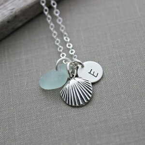 May include: A silver necklace with a seashell charm, a small piece of sea glass, and a silver charm with the letter 'E' on it.