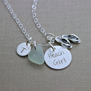 May include: Silver necklace with charms, including a sea glass charm, a charm with the letter T, a charm with the words "Beach Girl" and a charm of flip flops.