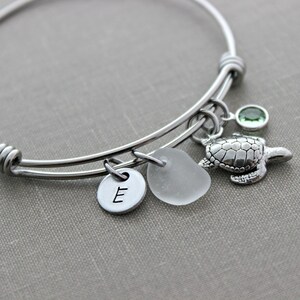 May include: Silver bangle bracelet with a charm of a sea turtle, a small piece of sea glass, and a letter 'E' charm. The bracelet also has a small green gemstone charm.