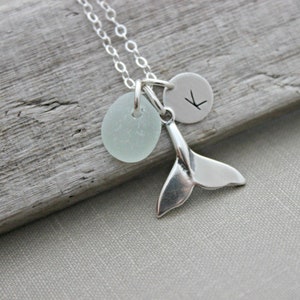 May include: A silver necklace with a whale tail charm, a small round charm with the letter "K" engraved on it, and a light blue sea glass charm.