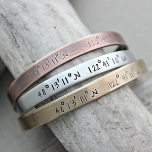 May include: Three stacked bracelets with coordinates engraved on them. The top bracelet is copper, the middle bracelet is silver, and the bottom bracelet is gold. The coordinates are 48°13′11″N 122°41' 10" W.