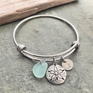 May include: A silver bangle bracelet with three charms: a light blue sea glass charm, a silver sand dollar charm, and a silver charm with the letter "J".