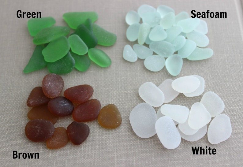 May include: Four piles of sea glass in different colors: green, seafoam, brown, and white. The sea glass is arranged in a square pattern on a light brown surface.