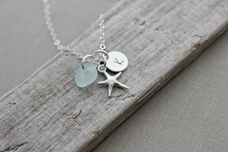 May include: A silver necklace with a light blue sea glass charm, a silver starfish charm, and a silver disc charm with the letter "K" engraved on it.