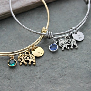 May include: Two gold and silver bangle bracelets with charms. The charms include an elephant, a letter, and a small gemstone. The bracelets are on a gray background.