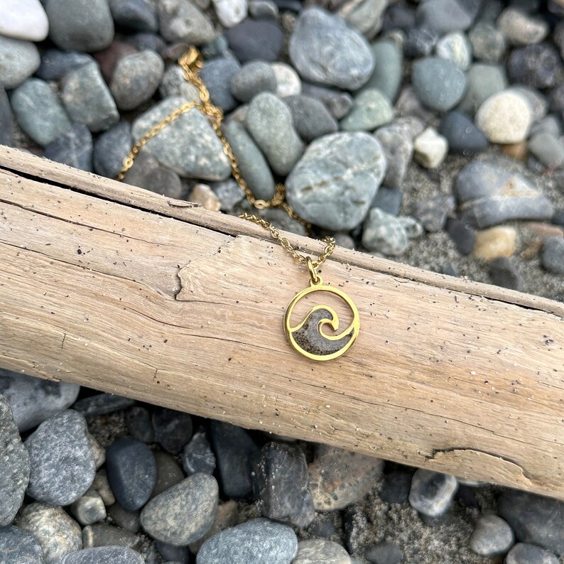 May include: A gold necklace with a round pendant featuring a wave design filled with sand. The necklace is on a wooden surface with small rocks.