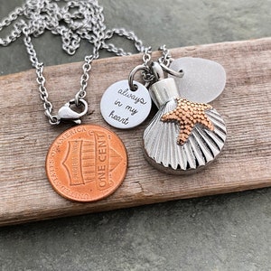 May include: Silver chain necklace with a seashell charm, a small round charm with the inscription "always in my heart", and a small glass bead. The necklace is lying on a piece of wood.